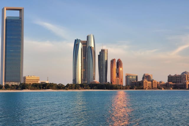 The Abu Dhabi skyline seen across the sea in low sunlight: a tall glass tower on the left, a cluster of curved glass towers in the centre and a domed, palace-like building on the right, behind a beach lined with palm trees.