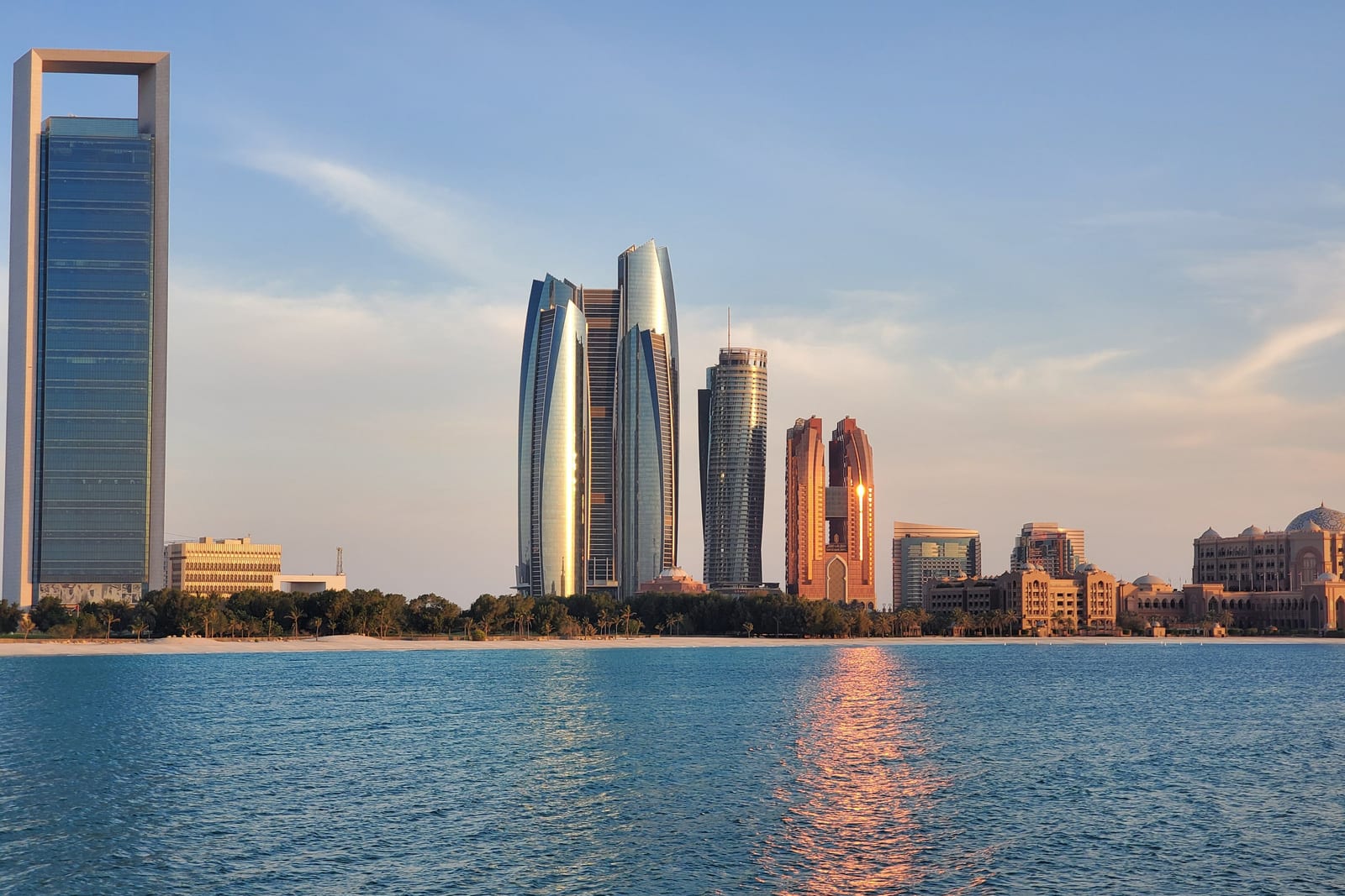 The Abu Dhabi skyline seen across the sea in low sunlight: a tall glass tower on the left, a cluster of curved glass towers in the centre and a domed, palace-like building on the right, behind a beach lined with palm trees.